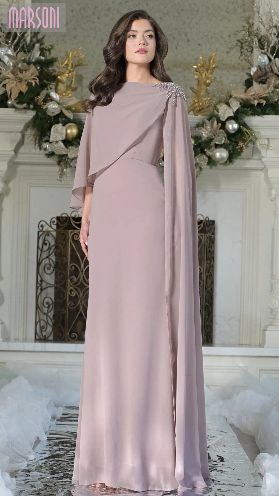 A woman in the Marsoni MV1362 long formal dress in dusty rose with cape sleeves stands indoors before a fireplace decorated with holiday ornaments.